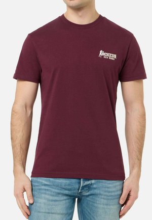 Mann trägt ein burgunderrotes T-Shirt mit dem Logo "Boxeur Des Rues" auf der Brust, kombiniert mit hellblauen Jeans, vor einfachem Hintergrund.