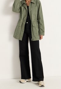 Chaqueta de color verde oliva con cuatro bolsillos frontales, cierre de botones y cremallera, combinada con pantalones de pierna ancha negros y zapatillas beige.