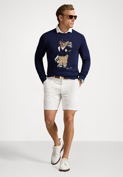 Il maglione blu navy presenta un orso ricamato in abbigliamento sportivo; abbinato a pantaloni corti bianchi e scarpe chiare, creando un look casual.