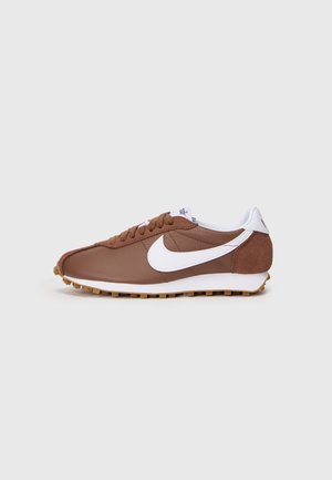 Bruine Nike hardloopschoen met wit swoosh-logo, vetersluiting, witte hielkraag en een gestructureerde gumzool op een effen grijze achtergrond.