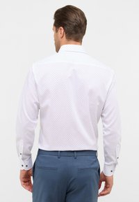 Witte shirt met kleine zwarte stippen, lange mouwen, knoopmanchetten en een gestructureerde kraag; gedragen met blauwe op maat gemaakte broek.