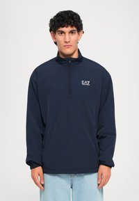 Muškarac nosi tamnoplavu jaknu s dugim rukavima i patentnim zatvaračem do četvrtine s EA7 Emporio Armani logom i svijetloplave traperice na bijeloj pozadini.