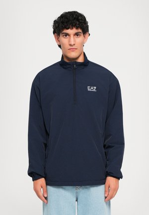 Man wearing navy blue long-sleeve quarter-zip jacket with EA7 Emporio Armani logo and light blue jeans against white background.