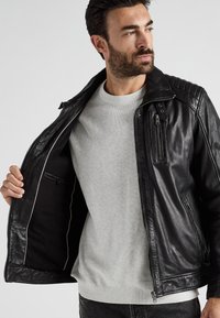 Svart läderjacka med ribbade axeldetaljer, zippered front och innerficka. Bärs över en ljusgrå tröja, strukturerad textil synlig.