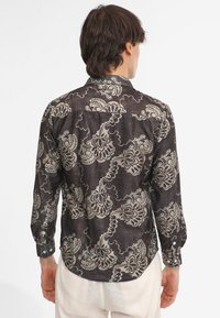 Maglietta nera con un motivo floreale in crema. Maniche lunghe, polsini con bottoni e orlo curvo. Realizzata in tessuto leggero con design testurizzato.