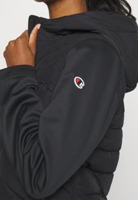 Czarna kurtka puchowa z kapturem, o gładkim wykończeniu, pikowanej strukturze i logo na rękawie. Posiada wysoki kołnierz oraz zapięcie na zamek.