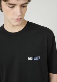 T-shirt en coton noir avec un petit logo brodé multicolore sur la poitrine indiquant "Sex Pistols". Col rond.