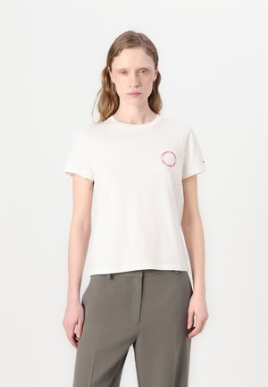 Cream-colored cotton t-shirt with short sleeves, round neckline, and circular red logo detail on the chest. Fitted design with smooth texture.