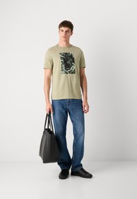 Oliven grønn bomulls t-skjorte med et svart og grønt grafisk trykk, kombinert med blå jeans og en svart lærveske. Svart joggesko fullfører stilen.