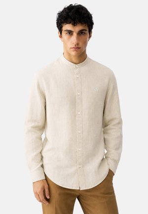 Giovane uomo con capelli ricci scuri che indossa una camicia beige con collo alla coreana e pantaloni marroni, in piedi con una mano in tasca.