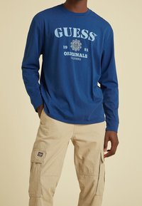 Langärmliges, blaues Baumwoll-T-Shirt mit "GUESS ORIGINALS" Grafik, kombiniert mit beigen Cargo-Hosen mit Seitentaschen.
