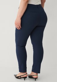 Marineblå tætsiddende bukser i et strækbart stof, med en mellem-høj talje, baglommer og et slankt, tapered design. Sorte hæle sko synlige.