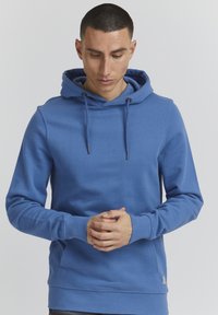 Sudadera de algodón azul con cordón ajustable, bolsillo canguro y puños acanalados. Textura suave y diseño ajustado, con una pequeña etiqueta de marca en el lateral.