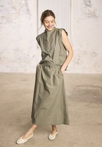 Femme souriante, portant un haut sans manches vert olive avec des épaules volantées et une longue jupe assortie, les mains dans les poches, debout sur un sol en béton.