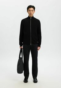 Juodas zip-up švarkas su aukštu apykakle, dėvimas ant tamsių džinsų. Modelis laiko tekstūruotą pilką rankinę, stovėdamas neutralioje fone.