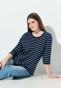 Tummansininen ja vaaleansininen raidallinen pitkähiha toppi, jossa pyöreä kaula-aukko, yhdistettynä vaaleansinisiin farkkuihin, yksinkertaista valkoista taustaa vasten.