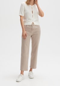 Mujer vestida con pantalones rectos de tiro alto en color beige, un cárdigan blanco de textura con mangas cortas y botones en la parte delantera, y zapatillas blancas con cordones sobre fondo blanco.