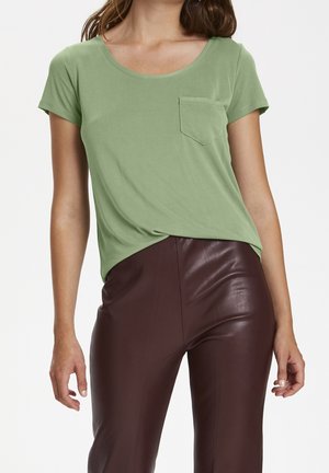 Kobieta ubrana w zieloną koszulkę z krótkim rękawem z kieszonką na klatce piersiowej oraz ciemnobrązowe skórzane spodnie z wysokim stanem, stojąca na jednolitym tle.