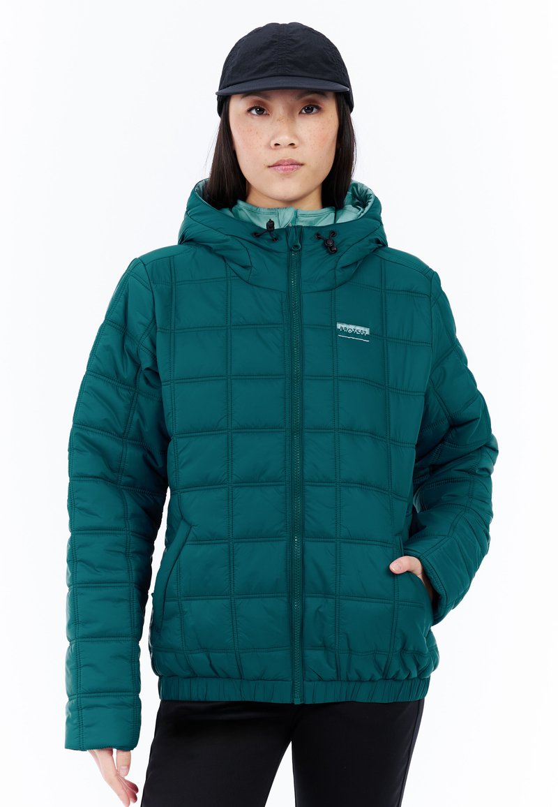 Vrouw in een teal quilted capuchonjack met rits, zwarte pet en zwarte broek, staand met één hand in de zak tegen een witte achtergrond.