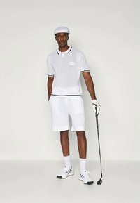 Λευκό golf κοντομάνικο μπλουζάκι με ριγωτή υφή, μαύρες λεπτομέρειες και λογότυπο. Συνδυασμένο με λευκά σορτς, καπέλο και γάντι του γκολφ, κρατώντας ένα μπαστούνι του γκολφ.