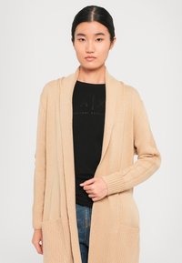Nuori nainen, jolla on päällään beigenvärinen neulecardigan mustan Armani Exchange -paidan ja sinisten farkkujen päällä, seisoo yksinkertaisen taustan edessä.