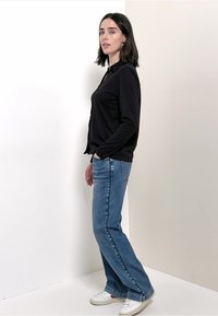 Zwarte lange mouwen shirt gecombineerd met lichtblauwe jeans. Witte sneakers. Model poseert zijwaarts tegen een neutrale achtergrond.