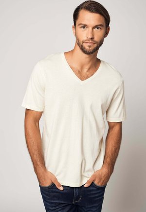 Maglietta a maniche corte e scollo a V color beige chiaro realizzata in tessuto morbido, caratterizzata da una vestibilità comoda e una texture liscia, abbinata a jeans blu scuro.