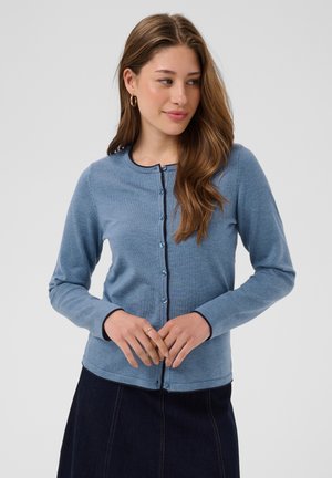 Femme aux longs cheveux bruns portant un cardigan bleu boutonné et une jupe sombre, debout, les mains jointes, regardant sur le côté devant un fond uni.