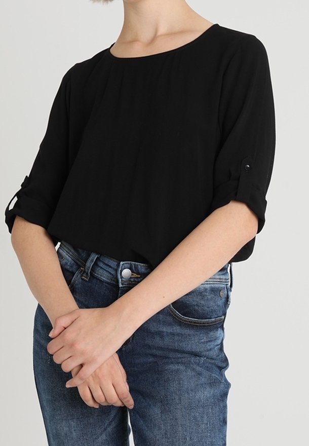 Person som bär en lös svart blus med uppvikta ärmar och blå denimjeans, stående med ihopknäppta händer framför sig.