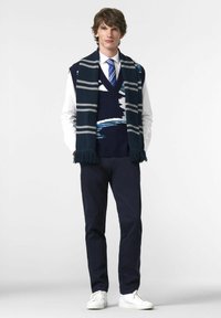 Navy knitted vest with abstract white and blue design, worn over a white shirt, paired with navy chinos and white sneakers, and a striped scarf.