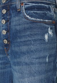 Jeans de mezclilla azul con cintura alta, que cuentan con un cierre de botón, una textura desgastada y patrones de desgaste visibles en la tela.