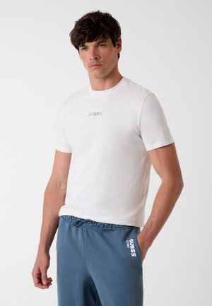 Man wearing white short-sleeve t-shirt and blue drawstring pants with "GUESS 1981" printed on left thigh, standing against white background.
