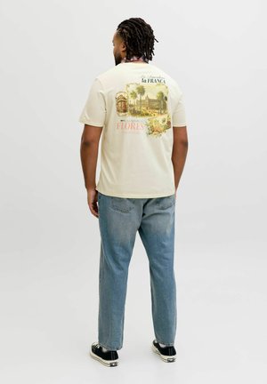 Hombre con el cabello trenzado de espaldas, lleva una camiseta crema con gráficos de ilustraciones vintage de ciudades españolas, jeans azules y zapatillas negras.