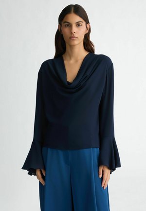 Maglietta a maniche lunghe blu navy con scollo drappeggiato e maniche svasate, abbinata a pantaloni larghi blu coordinati. Tessuto liscio, dettagli minimi.