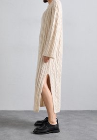 Kvinna som bär en beige kabelstickad lång klänning med sidoklyv och svarta läder snörskor, står på en grå golv.