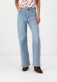 Jeans drepti de culoare albastru deschis, din denim, cu buzunare frontale, talie înaltă și margini tivite. Purtați cu botine maronii cu toc.