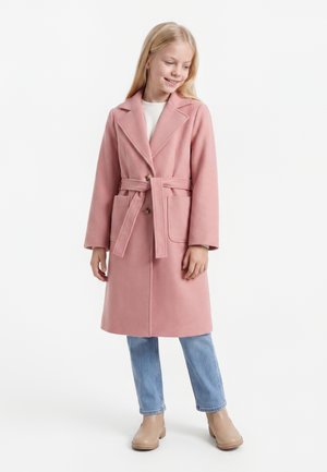Ung pige iført en pink frakke med bælte, lyseblå jeans og beige ankelstøvler, står og kigger nedad med et let smil.