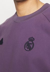 Sudadera morada con cuello redondo, que presenta un logo de corona texturizado y un emblema negro del Real Madrid en el pecho.