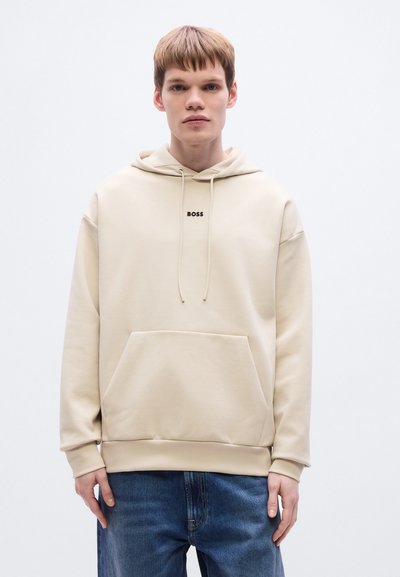 Sweat à capuche beige avec poche kangourou, capuche à cordon de serrage et logo noir "BOSS". Associé à un jean en denim bleu, fond neutre.