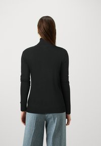 Femme avec de longs cheveux bruns portant un pull à col roulé noir et un jean large bleu clair, de dos contre un fond uni.