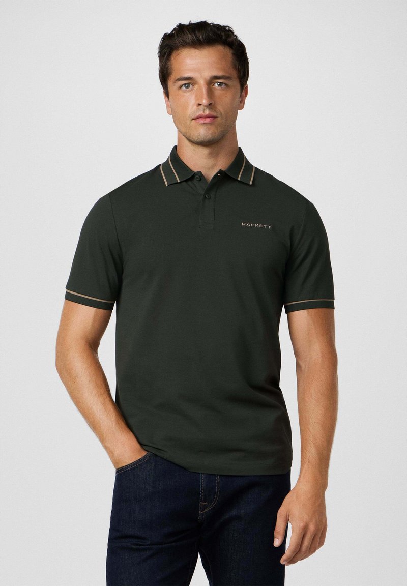 Man in een donkergroen polo shirt met een gestreepte kraag en mouwranden, staand met één hand in zijn zak tegen een effen achtergrond.