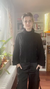 Črna pletena pulover s turtleneck ovratnikom in kabelskim vzorcem, v kombinaciji s črnimi širokimi hlačami. Notranji prostor z rastlinami in policami za knjige.
