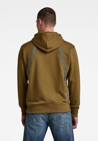 Sudadera marrón con capucha ajustable, espalda texturizada y acentos verdes en los hombros. Combina con jeans azules, que cuentan con un diseño clásico de cinco bolsillos.