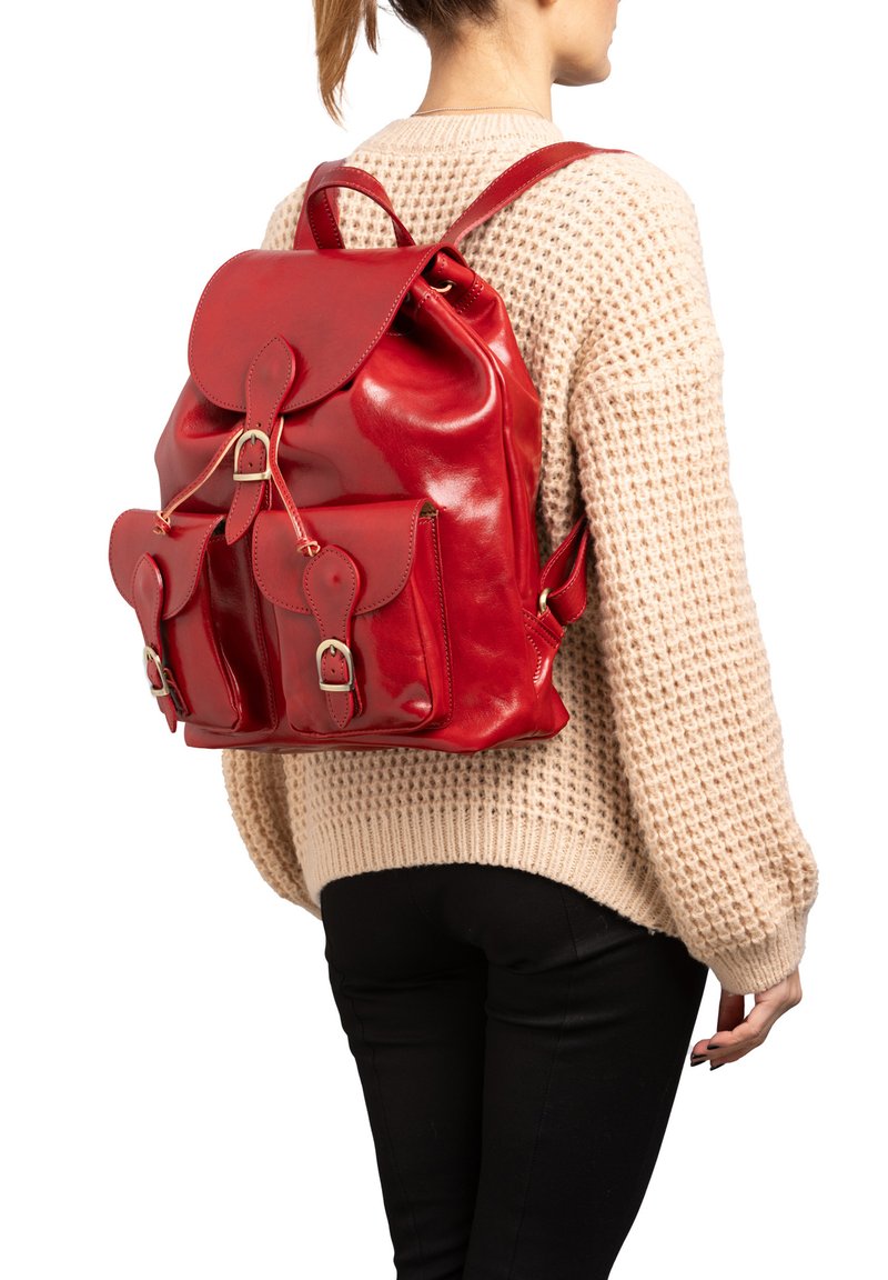 Persoon die een beige gebreide trui en een zwarte broek draagt, met een glanzende rode leren rugzak met voorzakken met gespen.