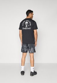 T-shirt noir à manches courtes avec le texte graphique blanc "ELEVATE YOUR GAINS" au dos. Associé à un short gris tie-dye et des baskets noires.