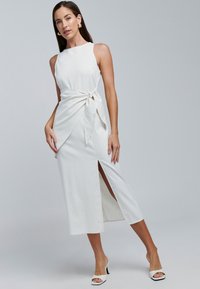 Witte linnenmix jurk met een nauwsluitend lijfje, een gestrikte taille en een zijsplit. Gecombineerd met witte open-toe hakken. Soepele textuur.