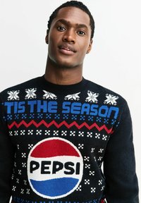 Schwarzer Pullover mit festlichen Mustern, "DIE JAHRESZEIT IST DA"-Text in Blau, Rot und weißen Akzenten, mit einem großen Pepsi-Logo in der Mitte.