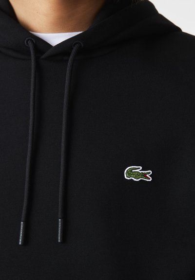 Haut à capuche noir avec cordons de serrage, arborant un petit logo de crocodile vert. Le tissu semble lisse et texturé, conçu avec une coupe classique.