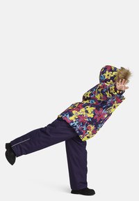 Färgglad pufferjacka med blommönster och huva med päls, kombinerad med mörklila byxor och svarta skor, stående i en lekfull pose.