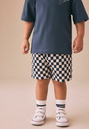 Barn iført marineblå t-shirt, sort-hvide ternede shorts, hvide sokker med sorte striber og hvide velcrolukninger-sneakers, står op.